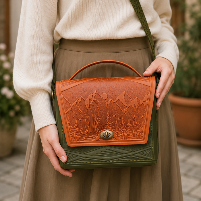 A woman wearing a cream ribbed sweater and a brown skirt carries a structured leather shoulder bag. The bag has a tan-brown embossed mountain and tree design on the front flap, a dark green base, and a matching green strap.