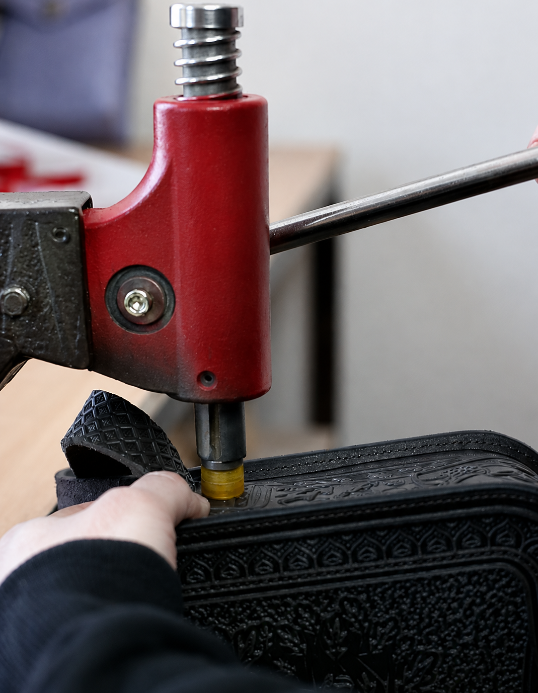 A close-up of a black embossed leather bag being processed by a red and silver leatherworking tool, with a person's hand holding the bag steady.